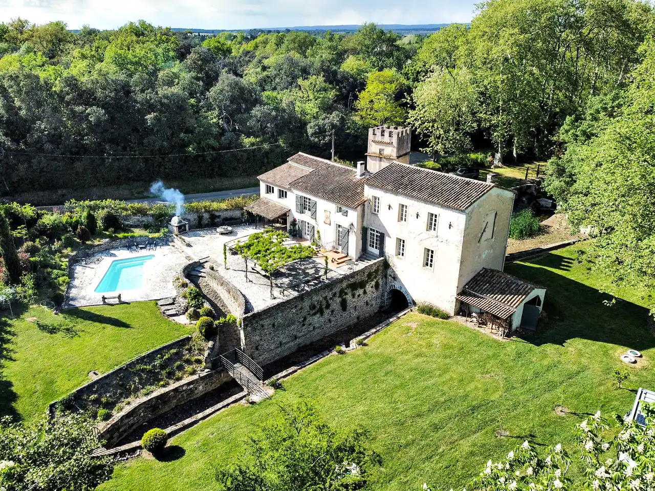 De Moulin d’Alzau van bovenaf: het huis met de toren, het zwembad op zijn terras, rook uit de pizzaoven, de pergola, de keermuur en de gazons, met bos rondom.